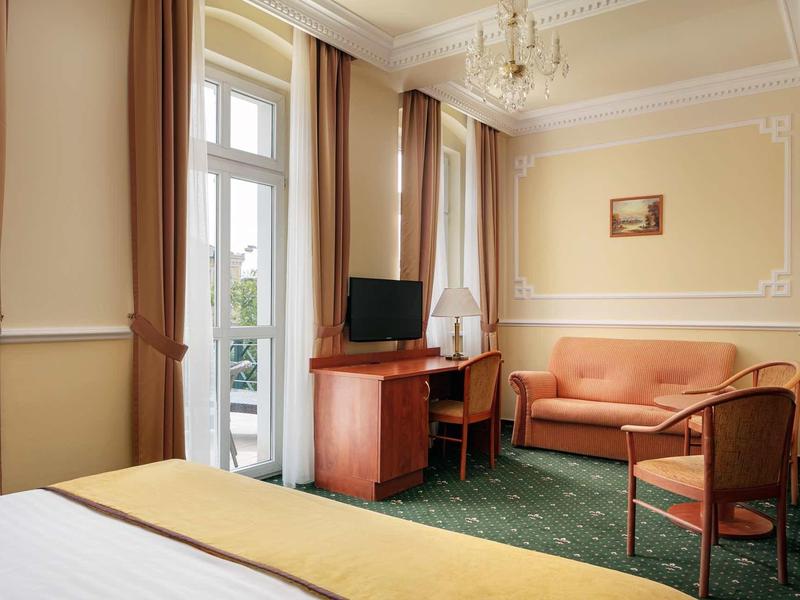 Bright hotel room with double bed, green carpet, wooden furniture, and a seating area.