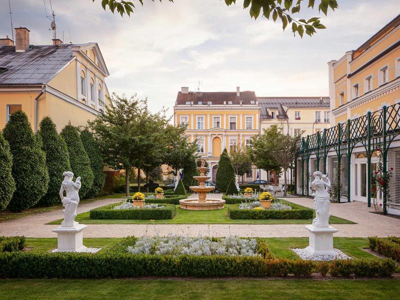 Elegant garden with fountain and statues between historic buildings at sunset.
