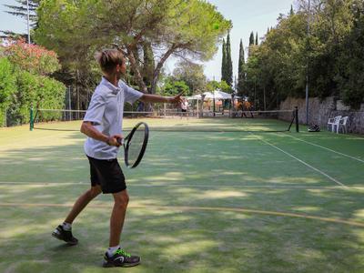 Junger Mann spielt Tennis auf einem von Bäumen umgebenen Platz bei Sonnenschein.