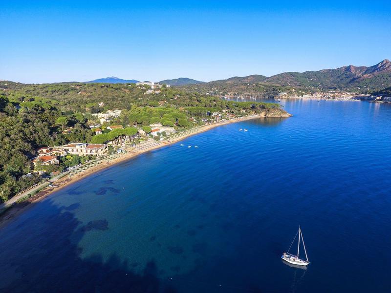 Küstenlandschaft mit Strand, Wald und Segelboot im klaren blauen Wasser unter klarem Himmel.