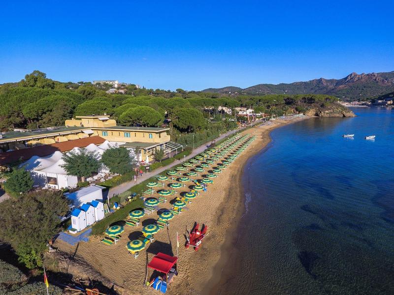Strand mit grünen Sonnenschirmen und Liegestühlen neben klarem blauen Wasser und bewaldetem Hügel im Hintergrund