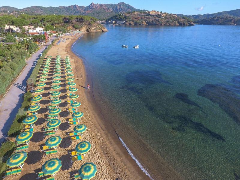 Strand mit grünen Sonnenschirmen, klarem Wasser und Hügeln im Hintergrund bei sonnigem Wetter.