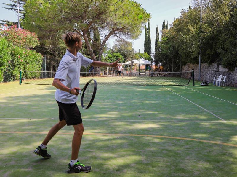 Junger Mann spielt Tennis auf einem Außenplatz, umgeben von Bäumen.