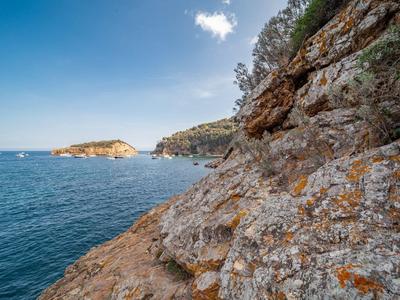 Küste mit felsigem Ufer, klarem blauem Meer und bewachsenen Hügeln unter blauem Himmel mit Wolken.