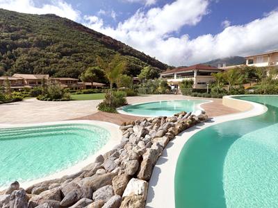 Kurviger Pool mit klarem Wasser, umgeben von Steinen, Häusern und bewaldetem Berg im Hintergrund.