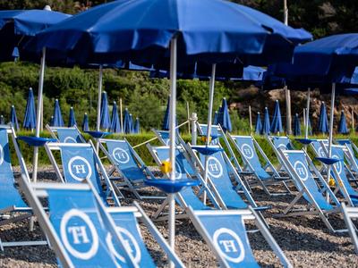Viele blaue Liegestühle mit weißen Symbolen und passenden blauen Sonnenschirmen an Kiesstrand.