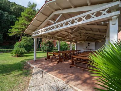 Überdachter Sitzbereich mit Holzbänken neben grünem Garten und gepflastertem Weg.