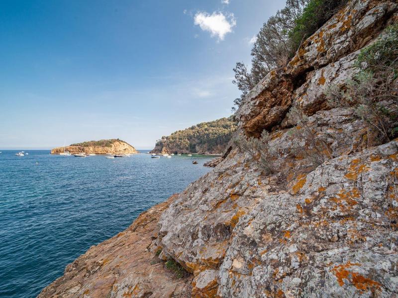 Küste mit felsigem Ufer, klarem blauem Meer und bewachsenen Hügeln unter blauem Himmel mit Wolken.