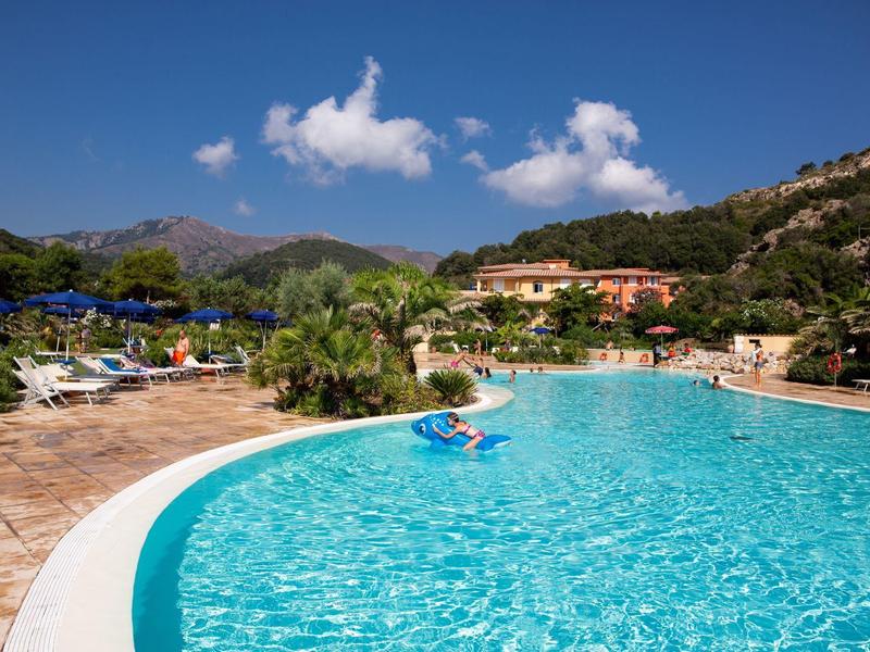 Großer blauer Pool mit Liegestühlen, Bergen und klarem Himmel im Hintergrund.