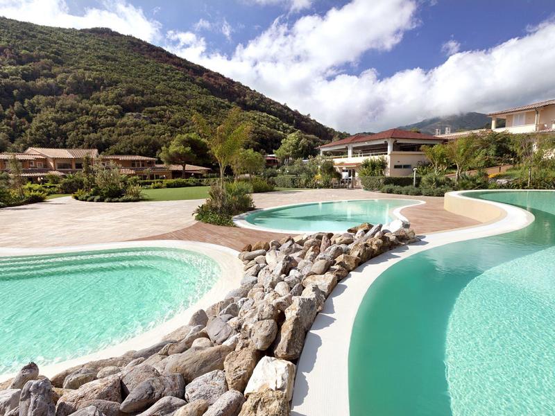 Kurviger Pool mit klarem Wasser, umgeben von Steinen, Häusern und bewaldetem Berg im Hintergrund.