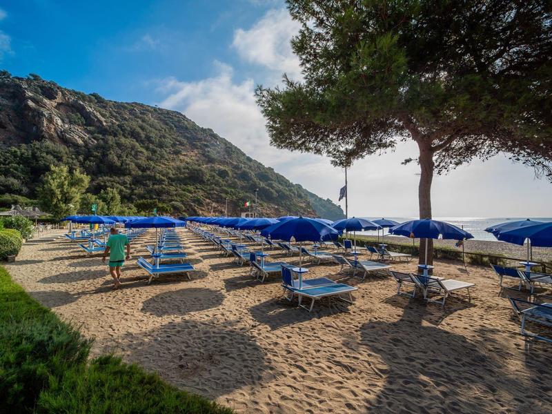 Sandstrand mit blauen Sonnenschirmen und Liegen, umgeben von grüner Vegetation und Hügeln im Hintergrund.