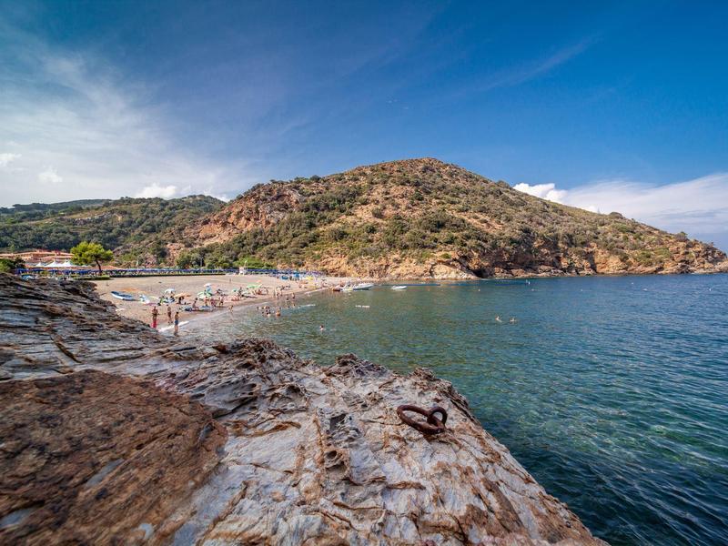 Klippen mit Felsen und ruhigem Meer, dahinter Strand mit Menschen und bewaldeten Hügeln unter blauem Himmel.