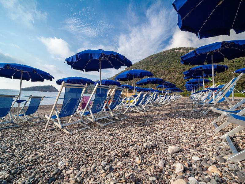 Strand mit Kieselsteinen, blauen Sonnenschirmen und Liegestühlen unter blauem Himmel.