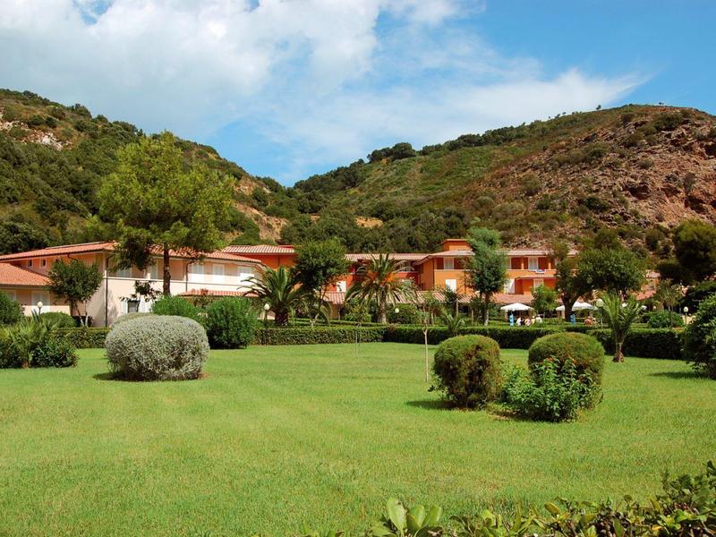 Grünes Gelände mit gepflegtem Rasen, Büschen, Palmen vor Gebäuden und Hügeln unter blauem Himmel.