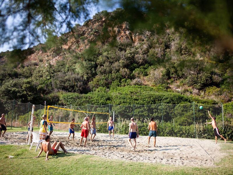 Menschen spielen Volleyball auf Sandplatz vor bewaldetem Hügel bei sonnigem Wetter.