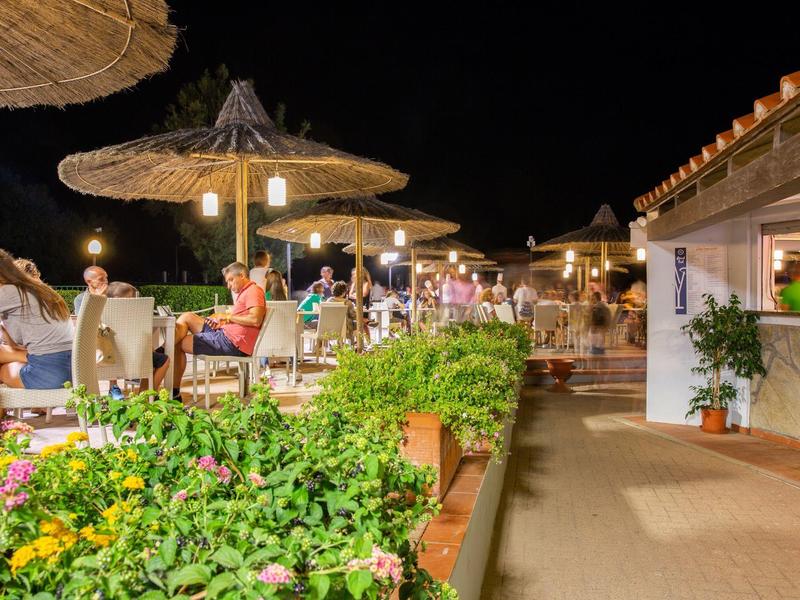 Beleuchtete Terrasse mit Gästen, grünen Pflanzen und Strohhut-Sonnenschirmen bei Nacht.