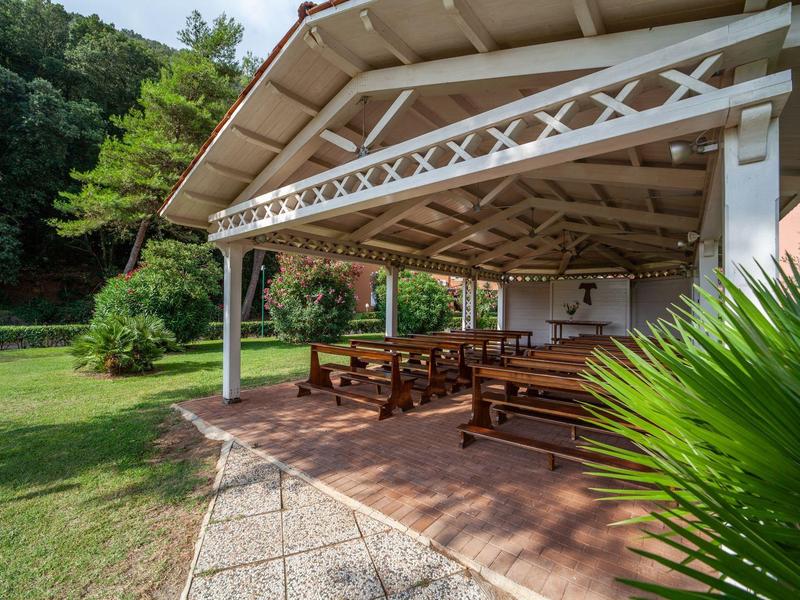 Überdachter Sitzbereich mit Holzbänken neben grünem Garten und gepflastertem Weg.