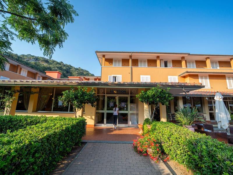 Mehrstöckiges gelbes Gebäude mit Veranda, grünen Hecken und sonnigem blauem Himmel.