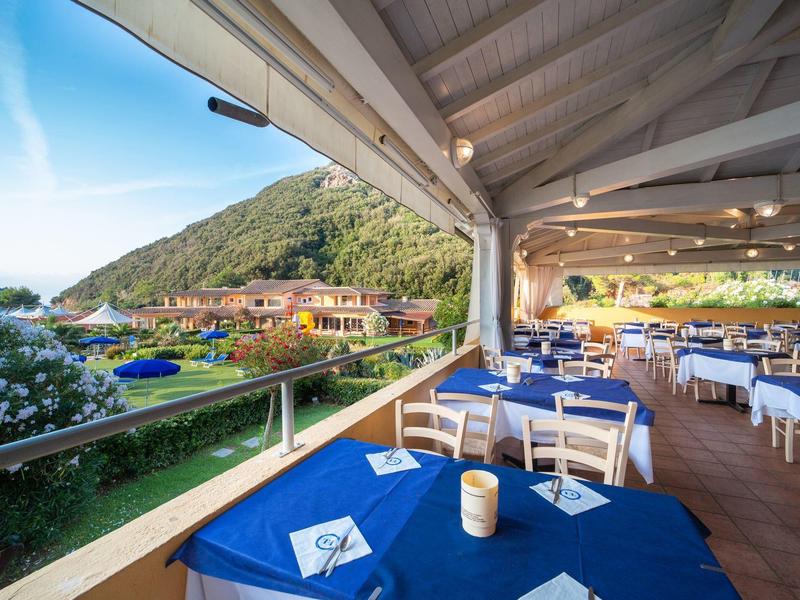 Überdachte Terrasse mit blauen Tischtüchern, weißen Stühlen und Blick auf grüne Hügel und Dorf.