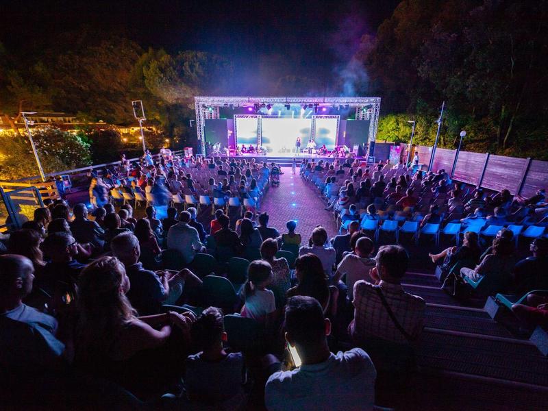Große Menschenmenge sitzt abends in Reihen vor einer beleuchteten Bühne im Freien.