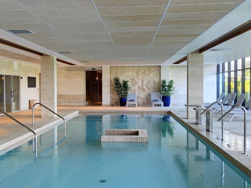 Binnenzwembad met ligstoelen en planten, omgeven door grote ramen.