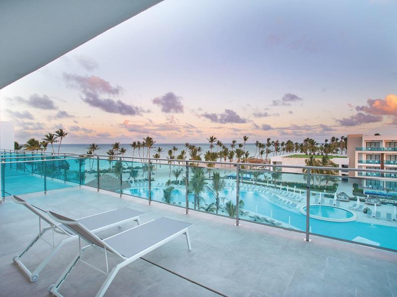 Balkon met stoel en glazen reling met uitzicht op een groot hotelzwembad en palmbomen bij zonsondergang.