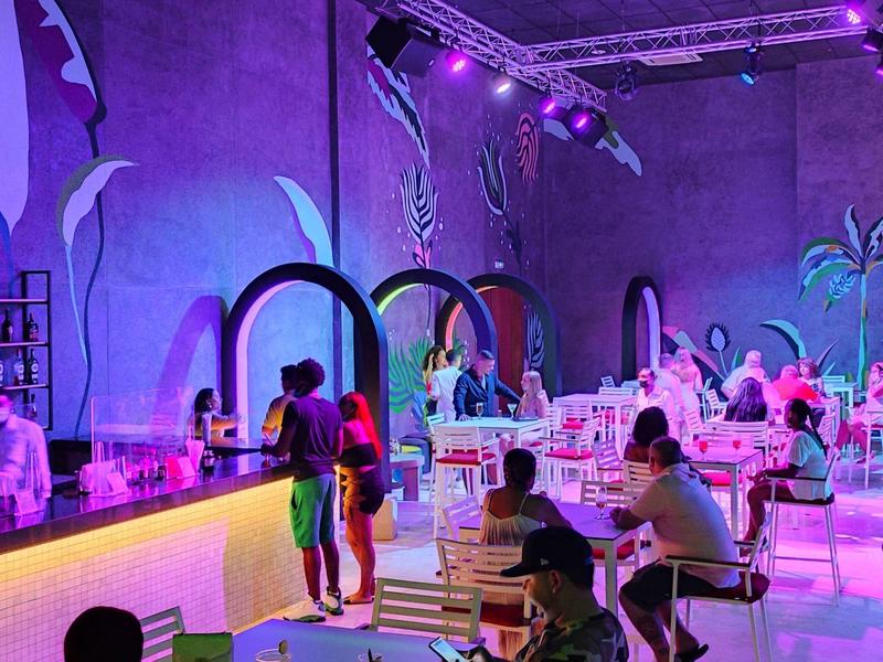 Moderne verlichte bar met gasten, kleurrijke lampen en tropische decoratie binnenin.