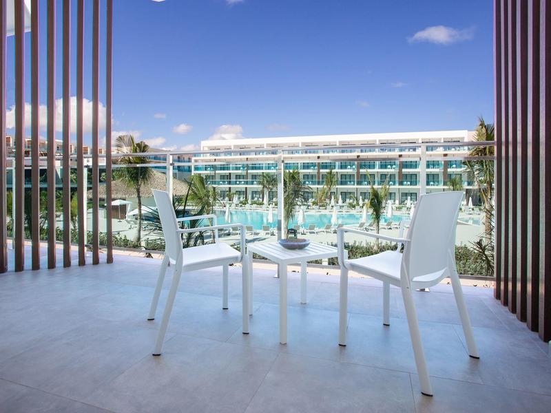 Balkon met witte tafel en stoelen met uitzicht op hotelzwembad en blauwe lucht.