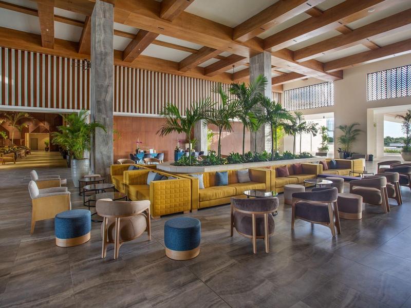 Grote hotel lobby met gele banken, houten tafels en planten in een moderne ruimte.