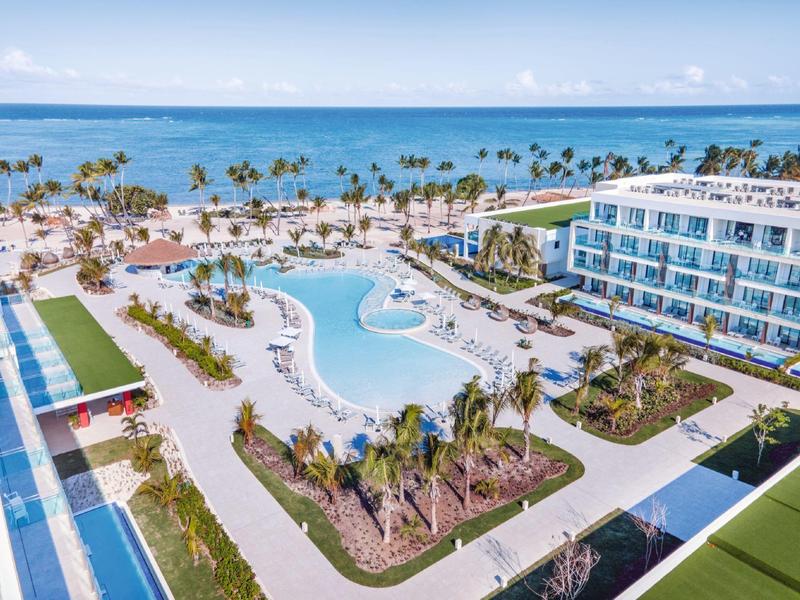 Uitzicht op een hotelresort met groot zwembad, palmbomen en zee op de achtergrond.