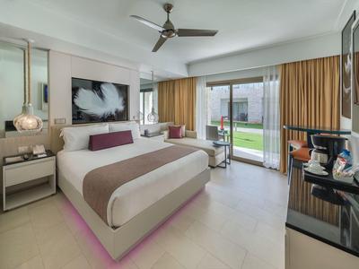 Helles Hotelzimmer mit großem Bett, Ventilator an der Decke und Blick auf Garten durch Glasschiebetür.