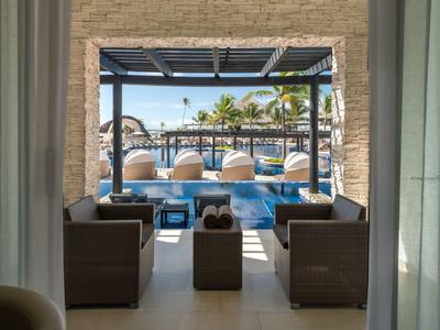 Blick aus einem schattigen Sitzbereich auf einen Pool mit Liegestühlen und Palmen.