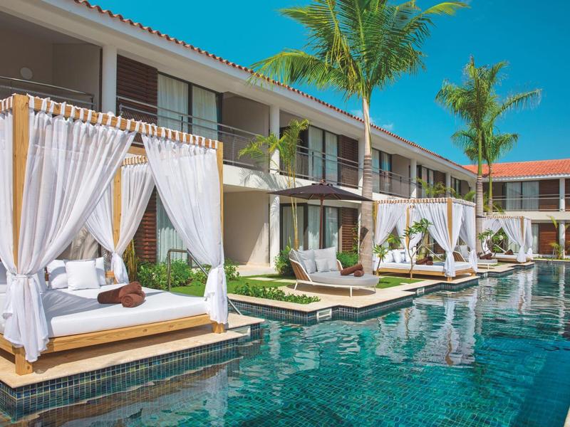 Piscina di un hotel moderno con tende bianche e palme tropicali sotto un cielo soleggiato.