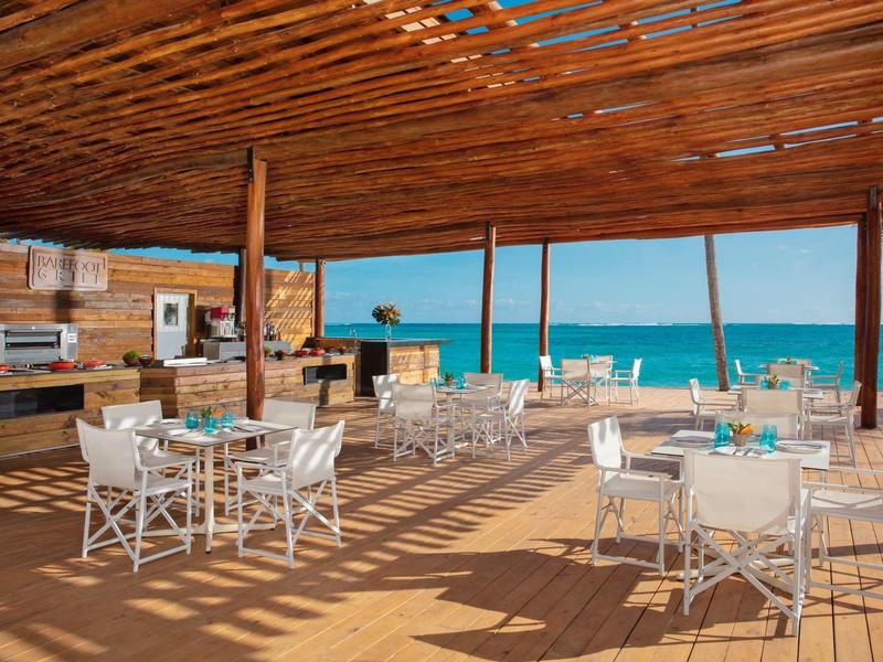 Ristorante sulla spiaggia aperto con sedie bianche e tetto in legno con vista sul mare.