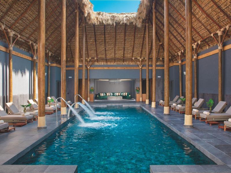 Elegante piscina interna con fontane d'acqua e lettini sotto un tetto di paglia.