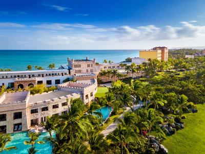 Hotel de lujo junto al mar con palmeras, piscina y jardín amplio bajo un cielo azul