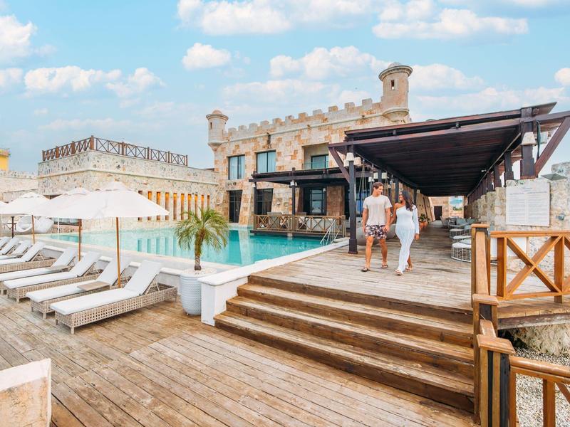 Hotelterras met zwembad, ligstoelen en mensen onder een blauwe lucht.