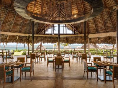 Offener, kreisförmiger Speisesaal unter Strohdach mit Blick auf Wasser und Palmen.