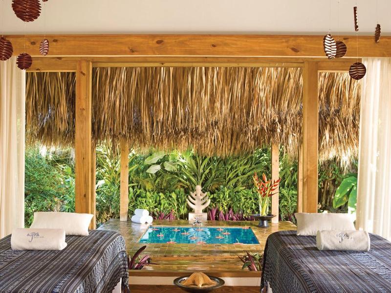 Zwei Betten mit dunklen Decken auf einer Veranda mit Blick auf einen Pool und tropische Pflanzen.