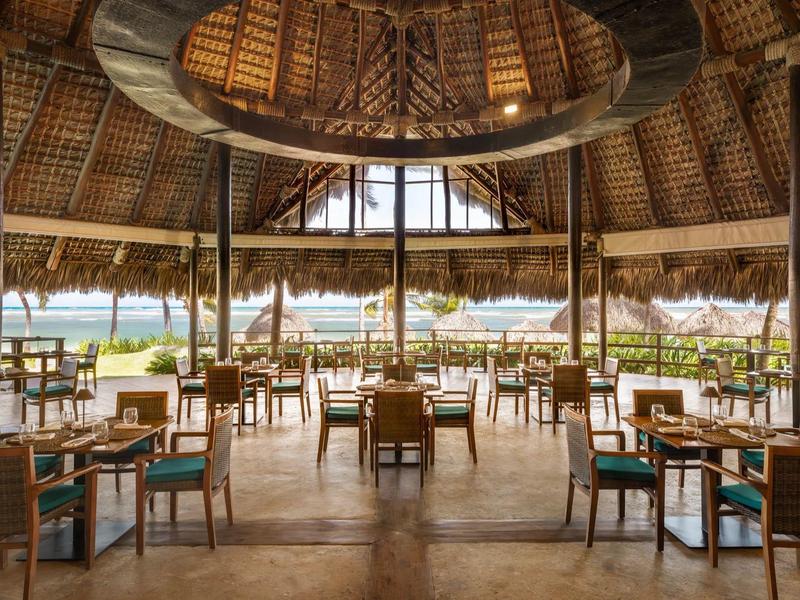 Offener, kreisförmiger Speisesaal unter Strohdach mit Blick auf Wasser und Palmen.