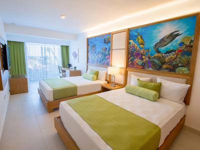 Hell eingerichtetes Hotelzimmer mit zwei Betten, grünen Akzenten und bunten Meeresgemälden.