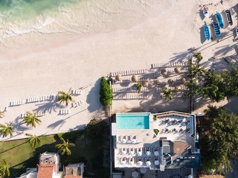 Luftaufnahme eines Strandes mit Sand, Palmen, Pool und blauem Meer am oberen Rand.