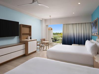 Bright hotel room with two beds, TV, dining table, and balcony access with a view.