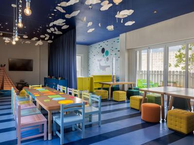 Colorful playroom with tables, chairs, carpet, and cloud design on ceiling in hotel.