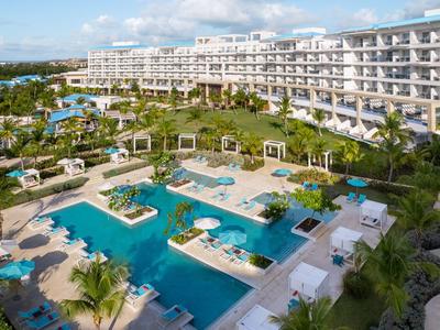 Large hotel complex with multiple pools, sun loungers, and Mediterranean garden under sunlight.