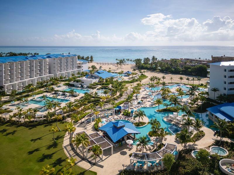Resort with multiple pools, palm trees, and ocean view under a partly cloudy sky.