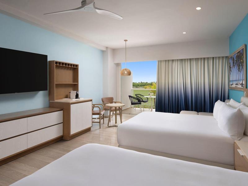 Bright hotel room with two beds, TV, dining table, and balcony access with a view.