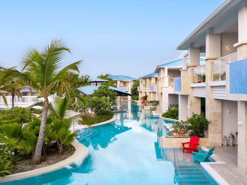 Modern hotel pool with palm trees and red chairs beside rooms under clear sky.