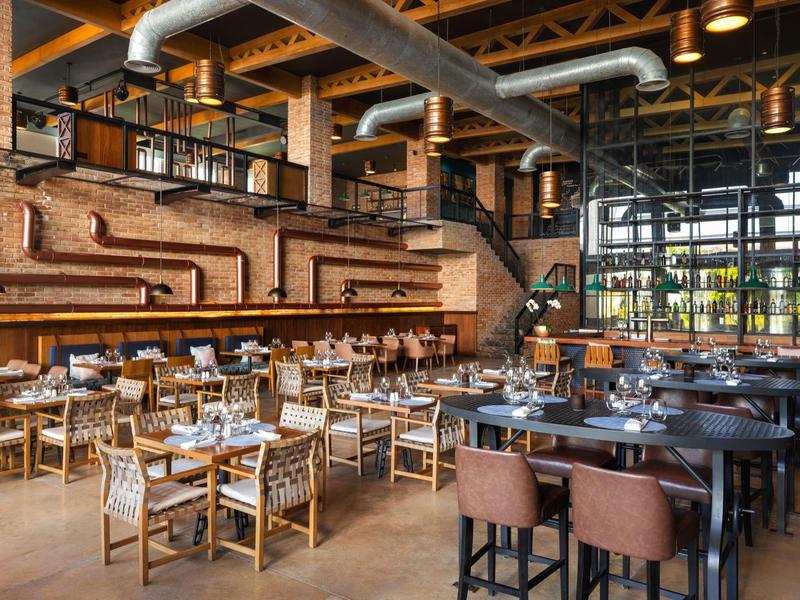 Modern restaurant with rustic interior, wooden tables, and leather chairs beneath exposed ceiling pipes.