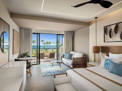 Modernes Hotelzimmer mit großem Bett, Sitzgruppe, TV, Balkon und Meerblick bei Tageslicht.